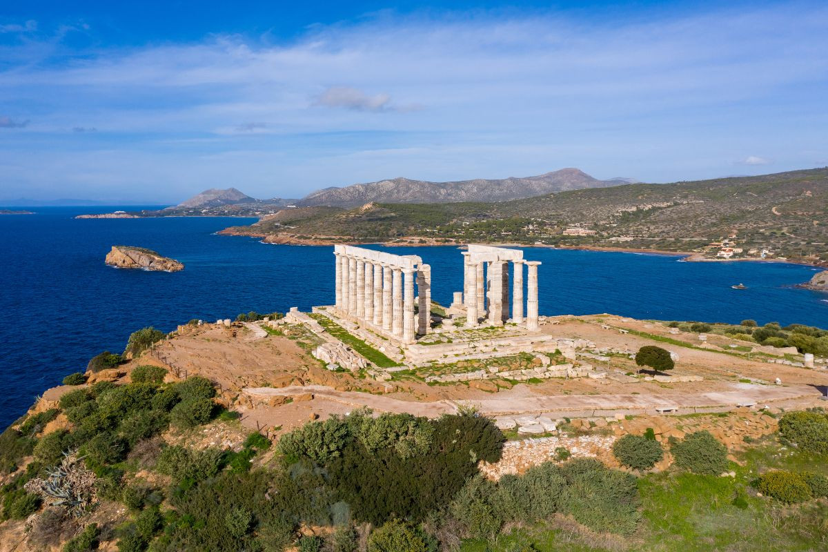 A scenic view of the Temple of Poseidon at Cape Sounio, perched on a cliff overlooking the deep blue Aegean Sea, with rolling hills in the background.